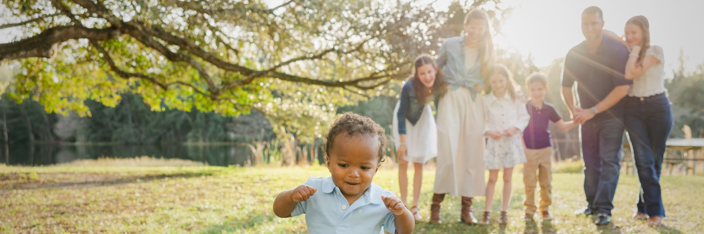 Small child with a family looking on outside on the grass with a lake in the background.